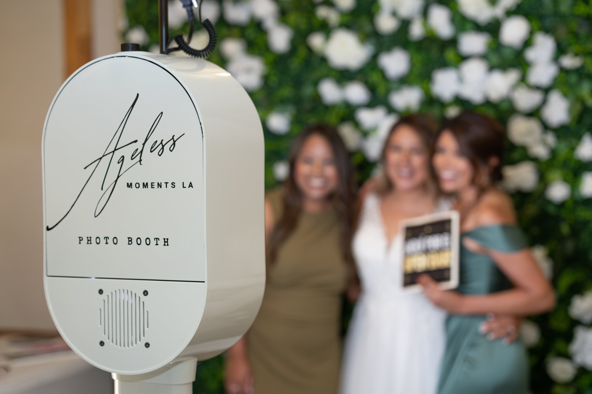 A photo booth unit in foreground, a group posing by floral wall
